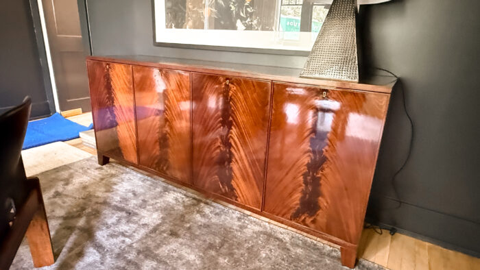 ustom high-gloss credenza with bookmatched wood grain doors, placed against a dark wall with framed art and a sculptural lamp on top.