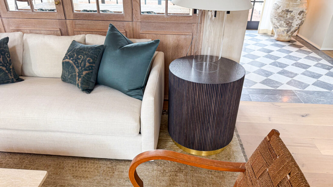 Round fluted wood side table with brushed brass base, placed beside a cream sofa with green accent pillows in a modern living space.