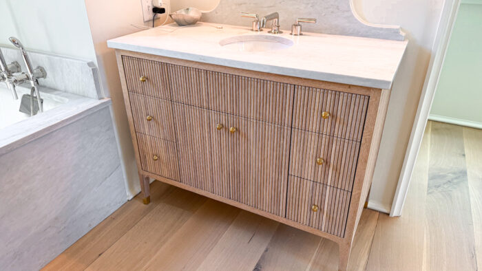 Custom fluted wood bathroom vanity with brass knobs and marble countertop, positioned beside a marble bathtub surround.