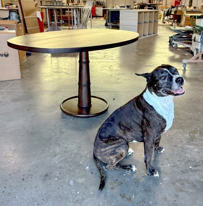 Round wooden dining table with dark walnut finish and single turned pedestal base standing in a woodworking workshop, with a brindle and white dog sitting nearby
